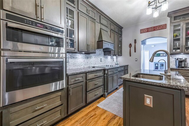 kitchen with light stone countertops, sink, stainless steel double oven, an island with sink, and light wood-type flooring