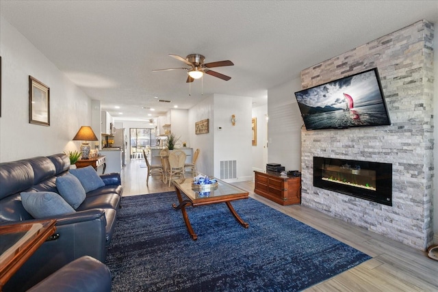 living room featuring a textured ceiling, light hardwood / wood-style flooring, ceiling fan, and a stone fireplace