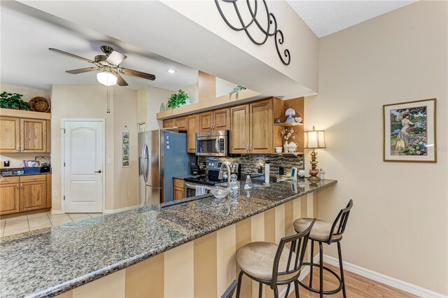 kitchen featuring a breakfast bar area, ceiling fan, dark stone countertops, kitchen peninsula, and stainless steel appliances