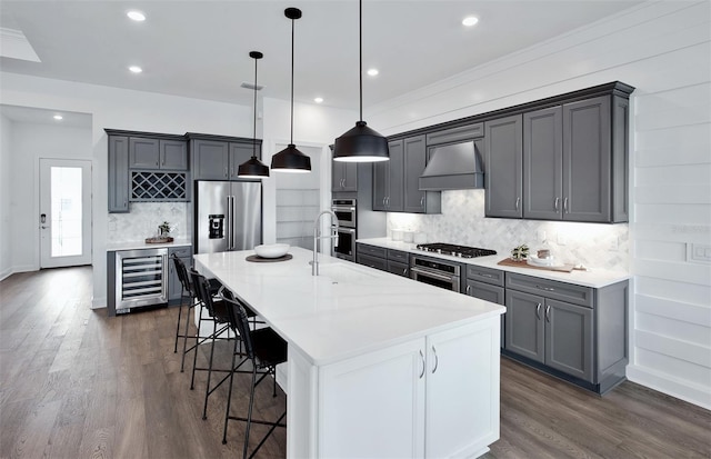 kitchen featuring pendant lighting, a kitchen island with sink, wine cooler, appliances with stainless steel finishes, and custom range hood