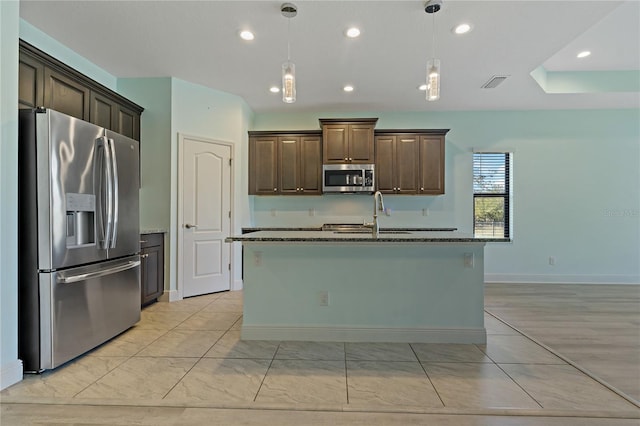 kitchen with appliances with stainless steel finishes, dark stone counters, dark brown cabinetry, pendant lighting, and a center island with sink