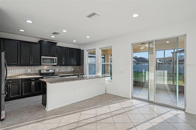 kitchen featuring a center island with sink, light tile patterned floors, appliances with stainless steel finishes, tasteful backsplash, and stone countertops