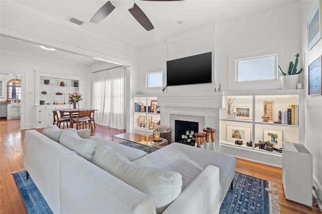 living room featuring light wood-type flooring, ceiling fan, crown molding, built in features, and a tiled fireplace