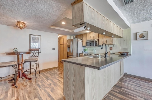 kitchen with kitchen peninsula, appliances with stainless steel finishes, light brown cabinetry, a textured ceiling, and hardwood / wood-style floors