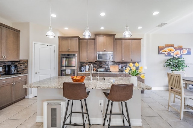 kitchen with light stone countertops, backsplash, an island with sink, decorative light fixtures, and appliances with stainless steel finishes