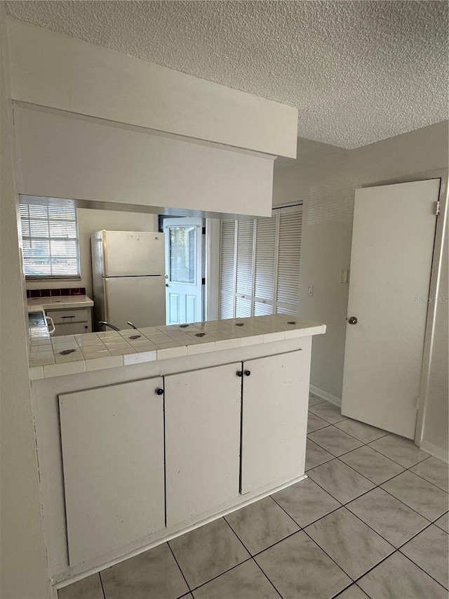 kitchen with light tile patterned flooring, kitchen peninsula, tile countertops, white fridge, and white cabinets