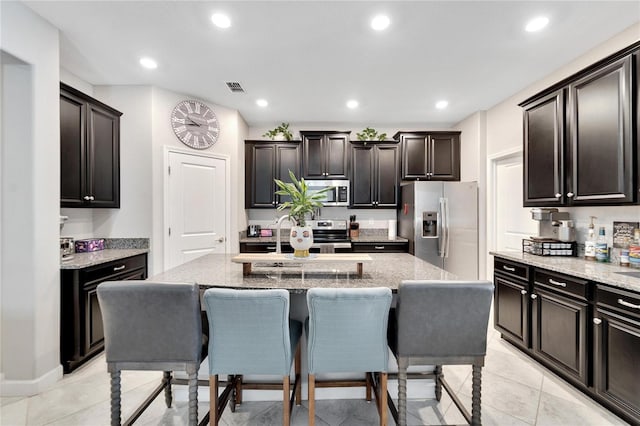 kitchen featuring appliances with stainless steel finishes, a center island with sink, light stone counters, and dark brown cabinets