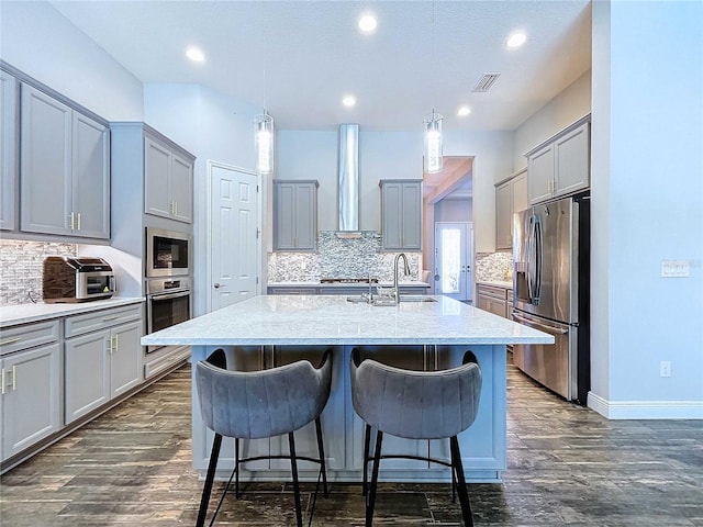 kitchen featuring stainless steel appliances, wall chimney range hood, an island with sink, decorative light fixtures, and gray cabinets