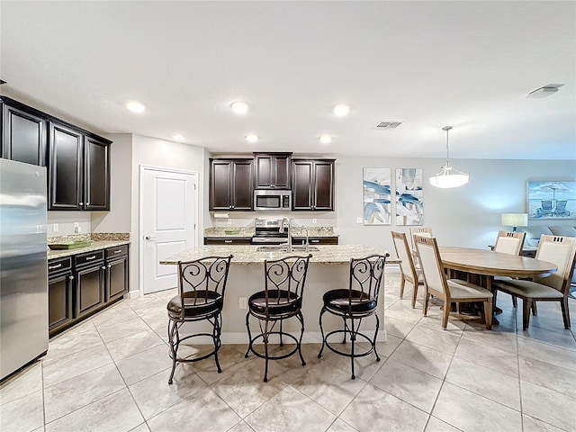 kitchen featuring a kitchen island with sink, appliances with stainless steel finishes, decorative light fixtures, light stone counters, and a breakfast bar area