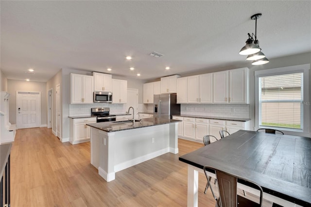 kitchen with dark stone counters, stainless steel appliances, decorative light fixtures, a center island with sink, and white cabinets