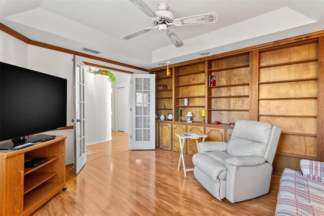 sitting room with french doors, light wood-type flooring, built in shelves, a raised ceiling, and ceiling fan