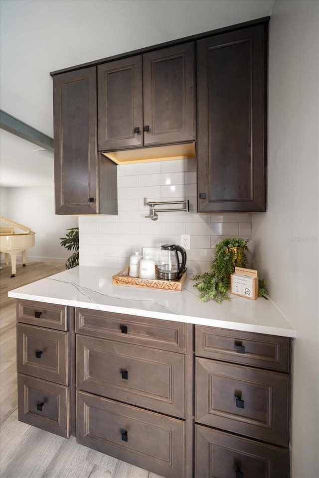 bar with decorative backsplash, light wood-type flooring, dark brown cabinetry, and light stone countertops