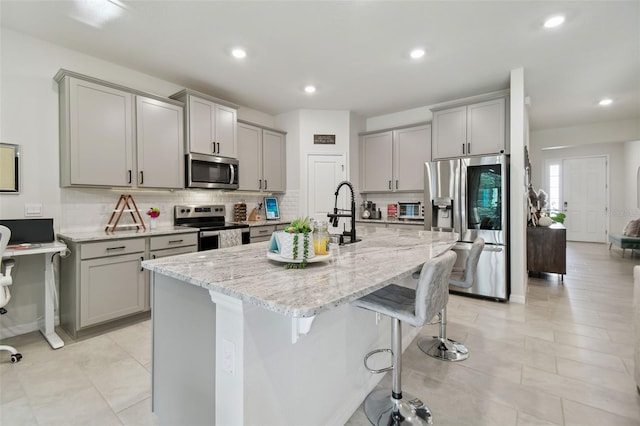 kitchen featuring gray cabinetry, light stone countertops, stainless steel appliances, and an island with sink