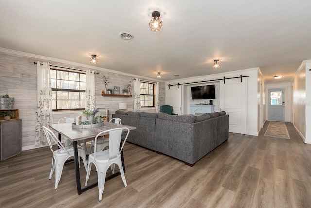 dining space featuring ornamental molding, a barn door, wooden walls, and dark wood-type flooring