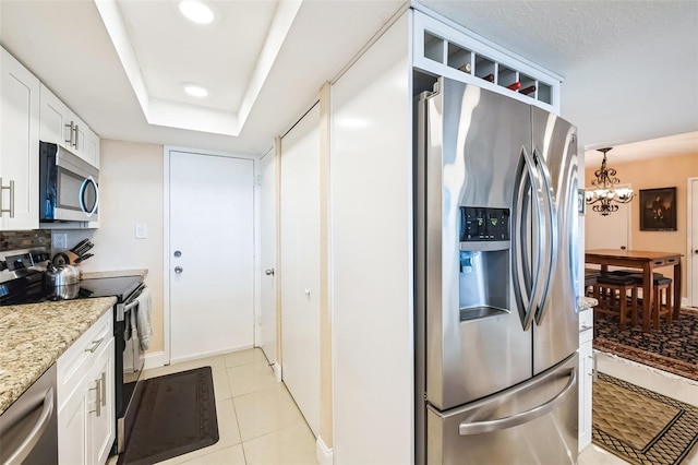 kitchen with light stone countertops, stainless steel appliances, a raised ceiling, a notable chandelier, and white cabinets