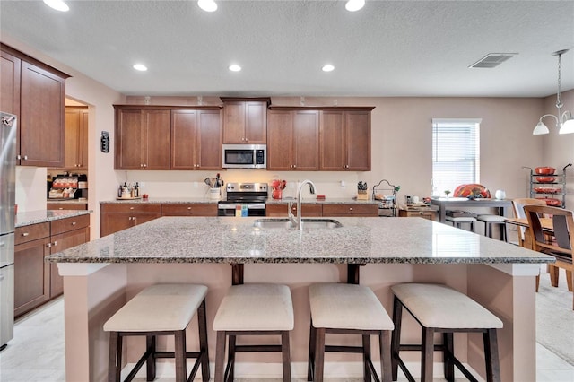 kitchen featuring light stone counters, stainless steel appliances, sink, a center island with sink, and hanging light fixtures