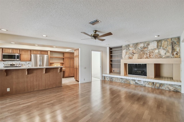 unfurnished living room with built in shelves, a textured ceiling, light wood-type flooring, ceiling fan, and a tiled fireplace