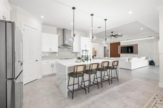 kitchen with wall chimney exhaust hood, a center island with sink, stainless steel refrigerator, pendant lighting, and white cabinets