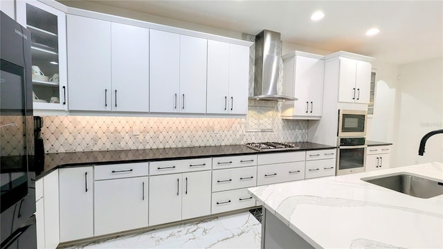 kitchen with appliances with stainless steel finishes, dark stone countertops, wall chimney range hood, white cabinets, and sink