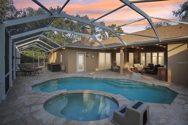 pool at dusk featuring an in ground hot tub, glass enclosure, and a patio area