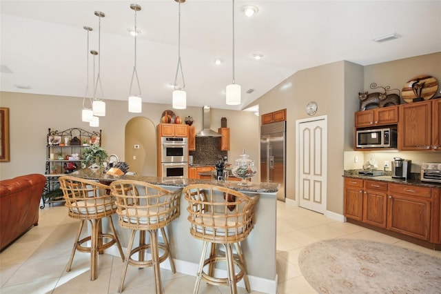 kitchen with hanging light fixtures, stainless steel appliances, wall chimney range hood, a kitchen bar, and light tile patterned floors