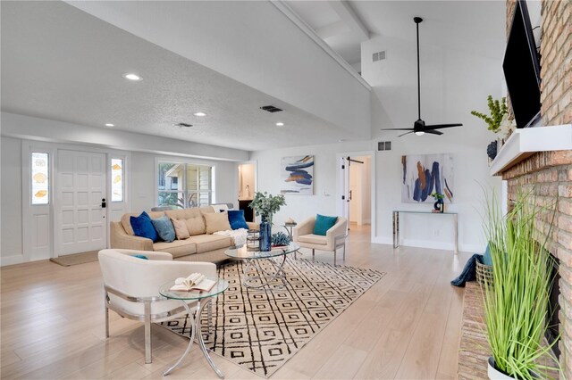 living room featuring a towering ceiling, a brick fireplace, a textured ceiling, ceiling fan, and light hardwood / wood-style flooring