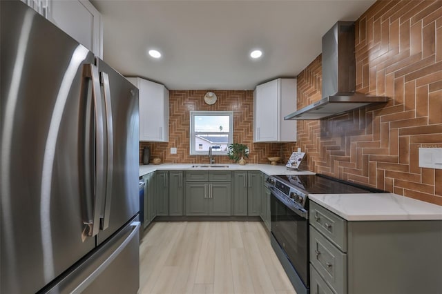 kitchen with black electric range oven, sink, wall chimney range hood, white cabinetry, and stainless steel refrigerator