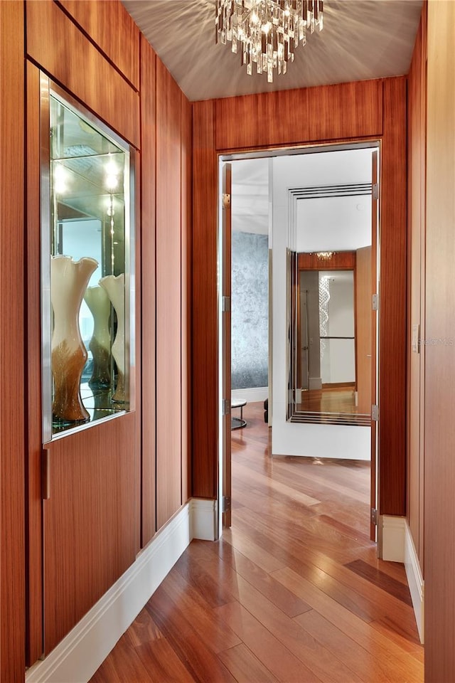 hallway with light wood-type flooring, an inviting chandelier, and wood walls