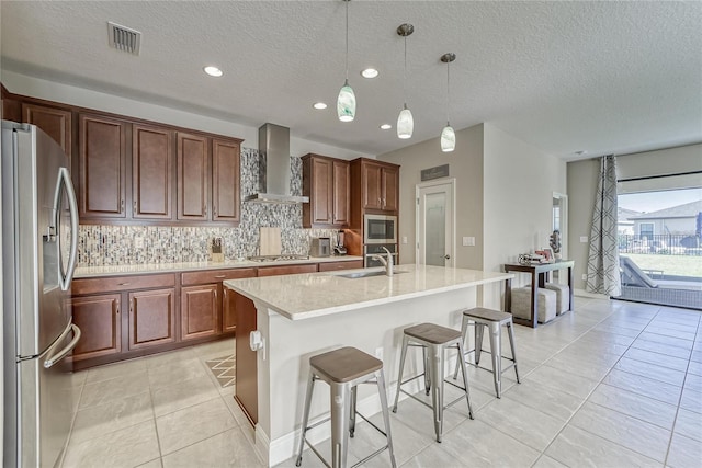 kitchen with wall chimney range hood, hanging light fixtures, sink, an island with sink, and appliances with stainless steel finishes