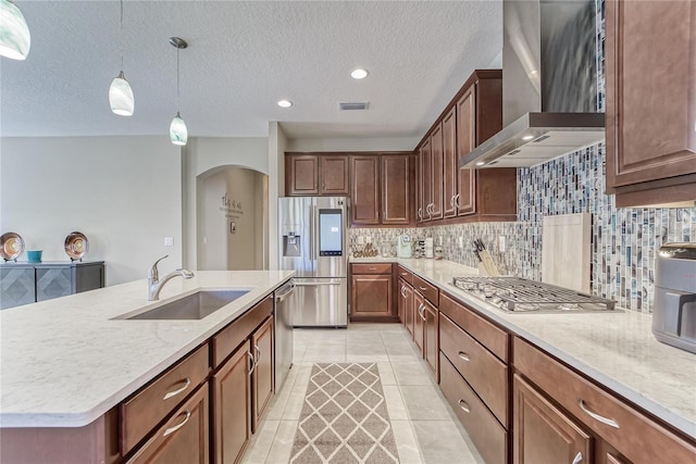 kitchen with pendant lighting, a kitchen island with sink, wall chimney range hood, sink, and stainless steel appliances