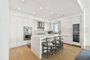 kitchen featuring double oven, a breakfast bar, white cabinets, and wall chimney range hood