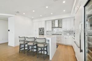 kitchen with a center island, wall chimney range hood, light hardwood / wood-style flooring, white cabinetry, and a breakfast bar area