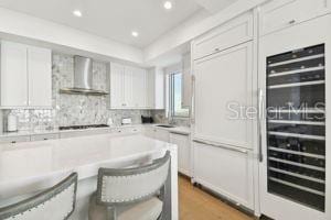 kitchen featuring wall chimney range hood, gas stovetop, a kitchen bar, white cabinetry, and beverage cooler