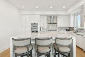 kitchen featuring stainless steel double oven, wall chimney range hood, a kitchen island, a breakfast bar area, and white cabinets