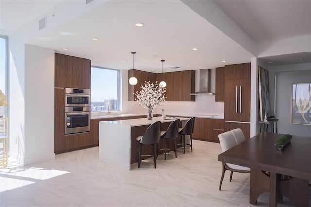 kitchen featuring decorative light fixtures, a breakfast bar area, stainless steel double oven, wall chimney range hood, and a center island with sink