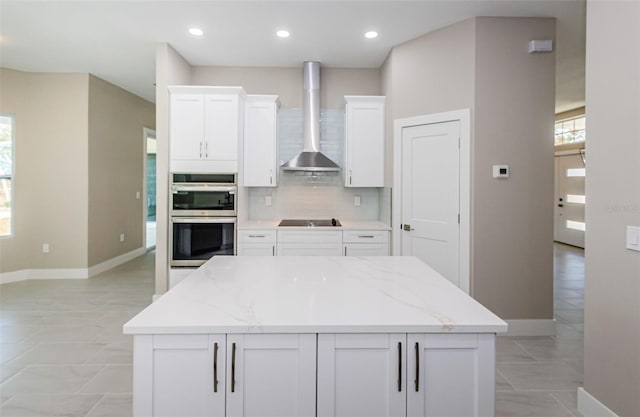 kitchen with double oven, white cabinetry, wall chimney exhaust hood, and light stone countertops