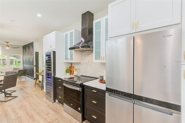 kitchen with stainless steel appliances, wall chimney range hood, light hardwood / wood-style floors, white cabinets, and ceiling fan