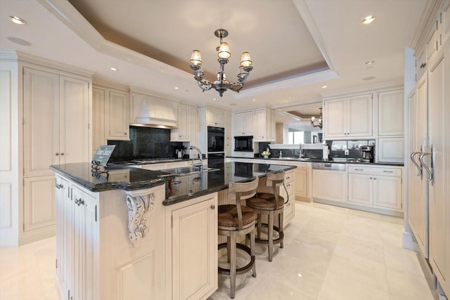 kitchen featuring premium range hood, a raised ceiling, a center island with sink, an inviting chandelier, and dishwasher