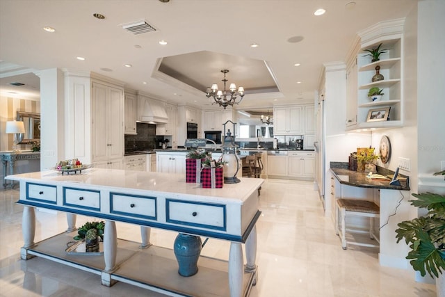 kitchen featuring visible vents, a raised ceiling, an island with sink, custom range hood, and an inviting chandelier