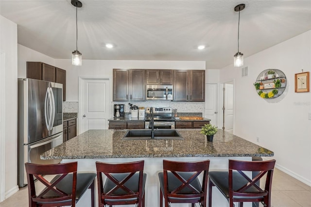 kitchen with a center island with sink, hanging light fixtures, dark stone countertops, dark brown cabinetry, and stainless steel appliances