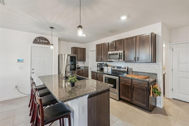 kitchen with appliances with stainless steel finishes, dark stone counters, sink, decorative light fixtures, and an island with sink