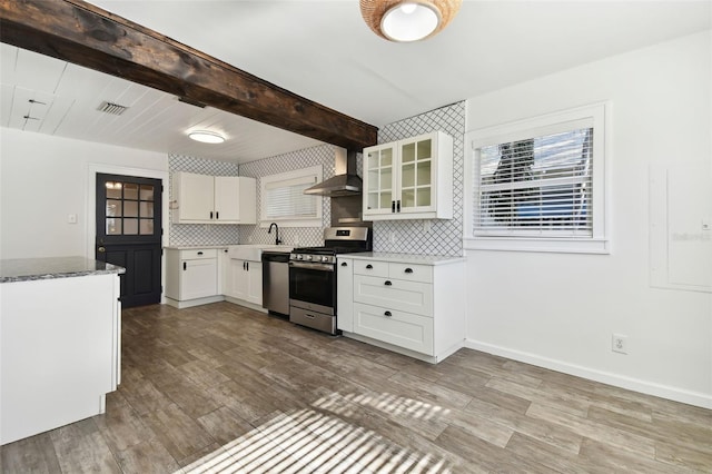 kitchen with appliances with stainless steel finishes, white cabinets, wall chimney exhaust hood, beamed ceiling, and light hardwood / wood-style floors