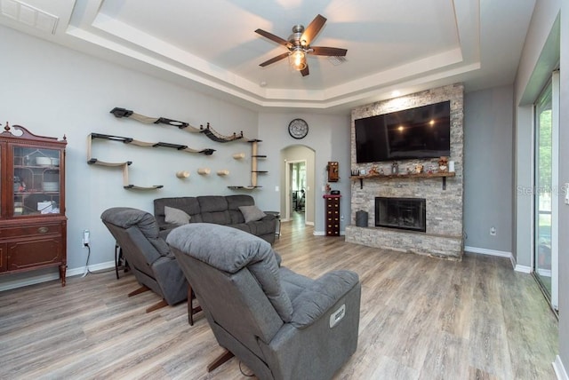 living room with ceiling fan, a stone fireplace, wood-type flooring, and a tray ceiling