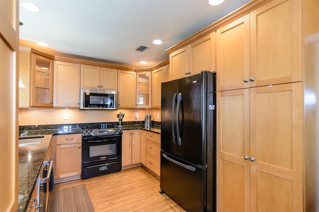 kitchen featuring light brown cabinets, light wood-type flooring, dark stone countertops, and black appliances