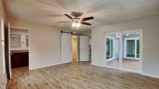 unfurnished bedroom with a textured ceiling, ceiling fan, a barn door, light hardwood / wood-style flooring, and connected bathroom