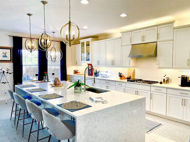 kitchen featuring a kitchen island with sink, a sink, decorative backsplash, stainless steel gas stovetop, and under cabinet range hood