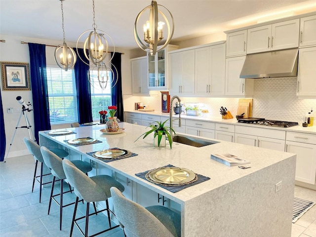 kitchen with under cabinet range hood, a center island with sink, stainless steel gas stovetop, white cabinetry, and a sink