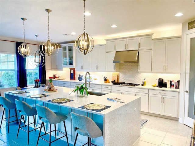 kitchen with an island with sink, under cabinet range hood, stainless steel gas stovetop, a breakfast bar area, and decorative backsplash