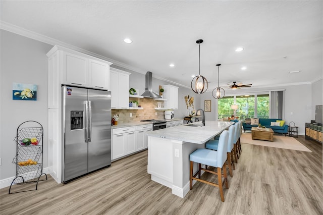 kitchen with white cabinets, a center island with sink, wall chimney range hood, ceiling fan, and stainless steel fridge