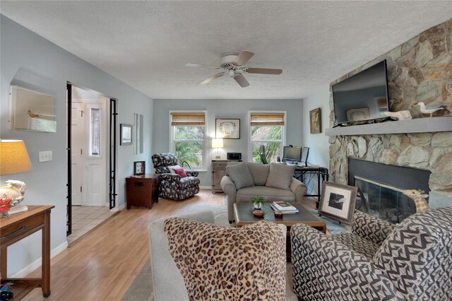 living room featuring a stone fireplace, ceiling fan, a textured ceiling, and light wood-type flooring